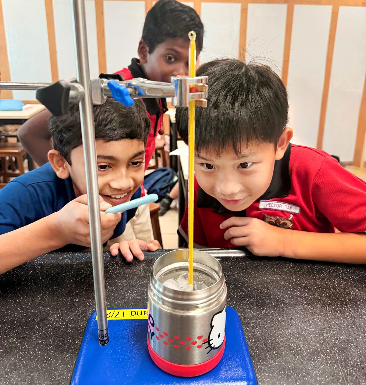 Four students in red shirts lean toward two microscopes on a classroom lab table.